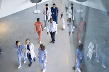 A large group of medical professionals are seen walking in a hall as they make their way to a medical conference.  They are each dressed professionally and are seen from above in this aerial view.