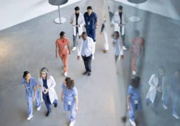 A large group of medical professionals are seen walking in a hall as they make their way to a medical conference.  They are each dressed professionally and are seen from above in this aerial view.
